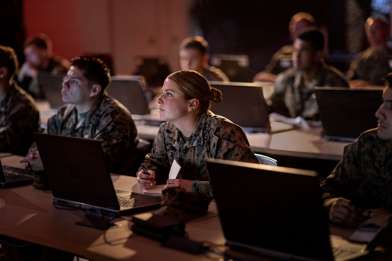 Marines in camouflage listen intently in a classroom setting, seated at desks with open laptops in front of them. The room is dimly lit. 