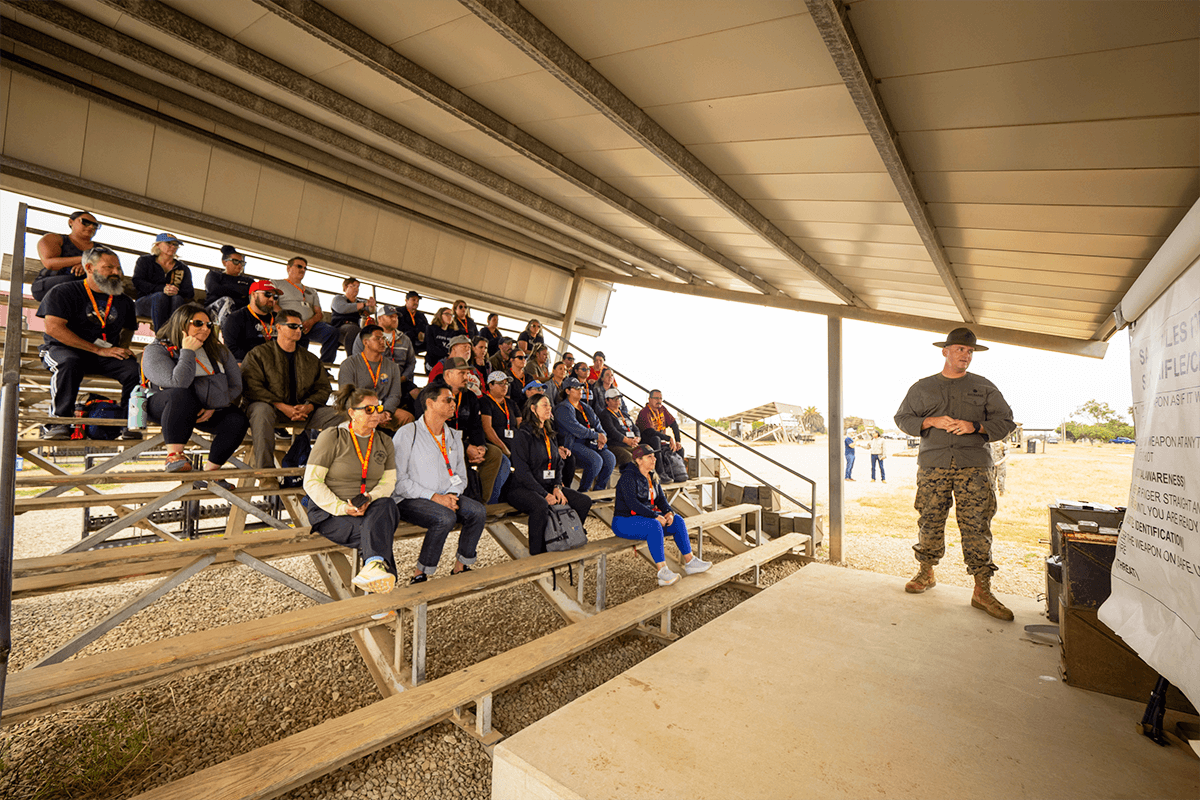A Marine Corps drill instructor wearing a campaign cover speaks to a large group of educators seated on bleachers.