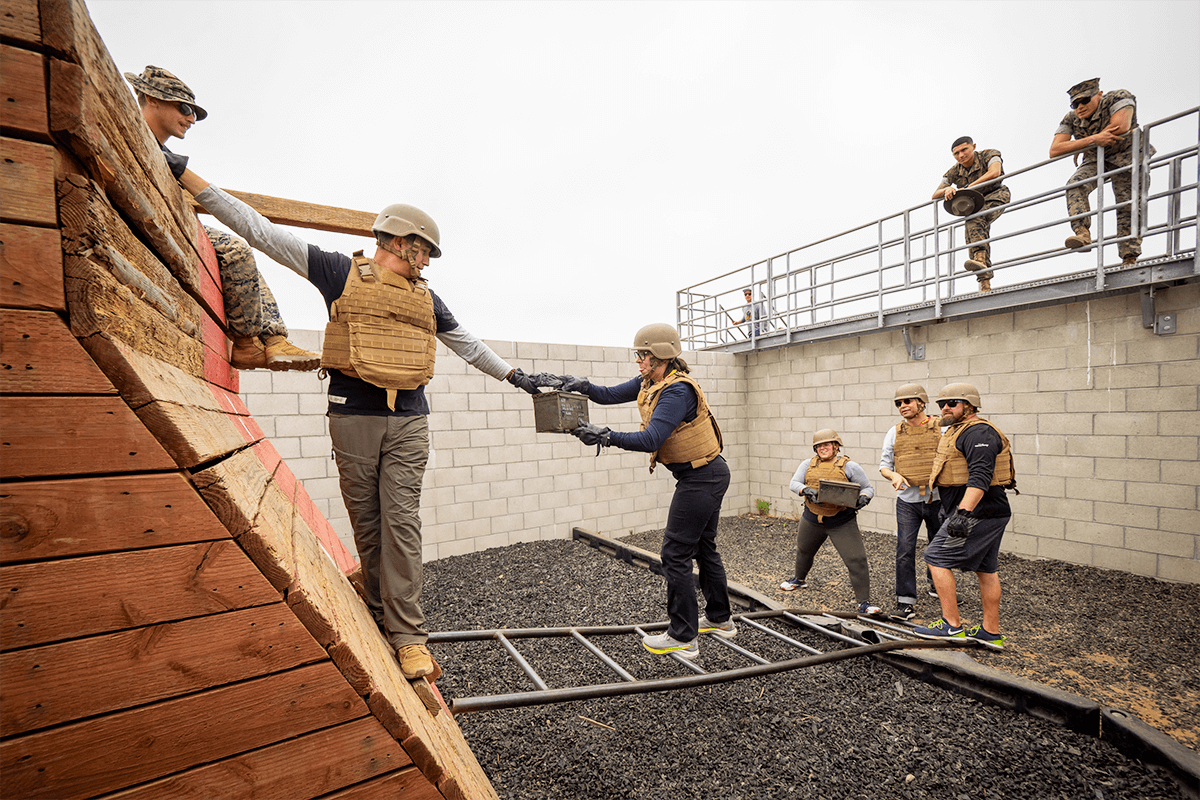 A male educator holding on to a wooden structure reaches for an ammo can from a female colleague standing on a ladder.
